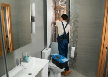 A man wearing overalls works on fixing a toilet, surrounded by plumbing tools and parts.